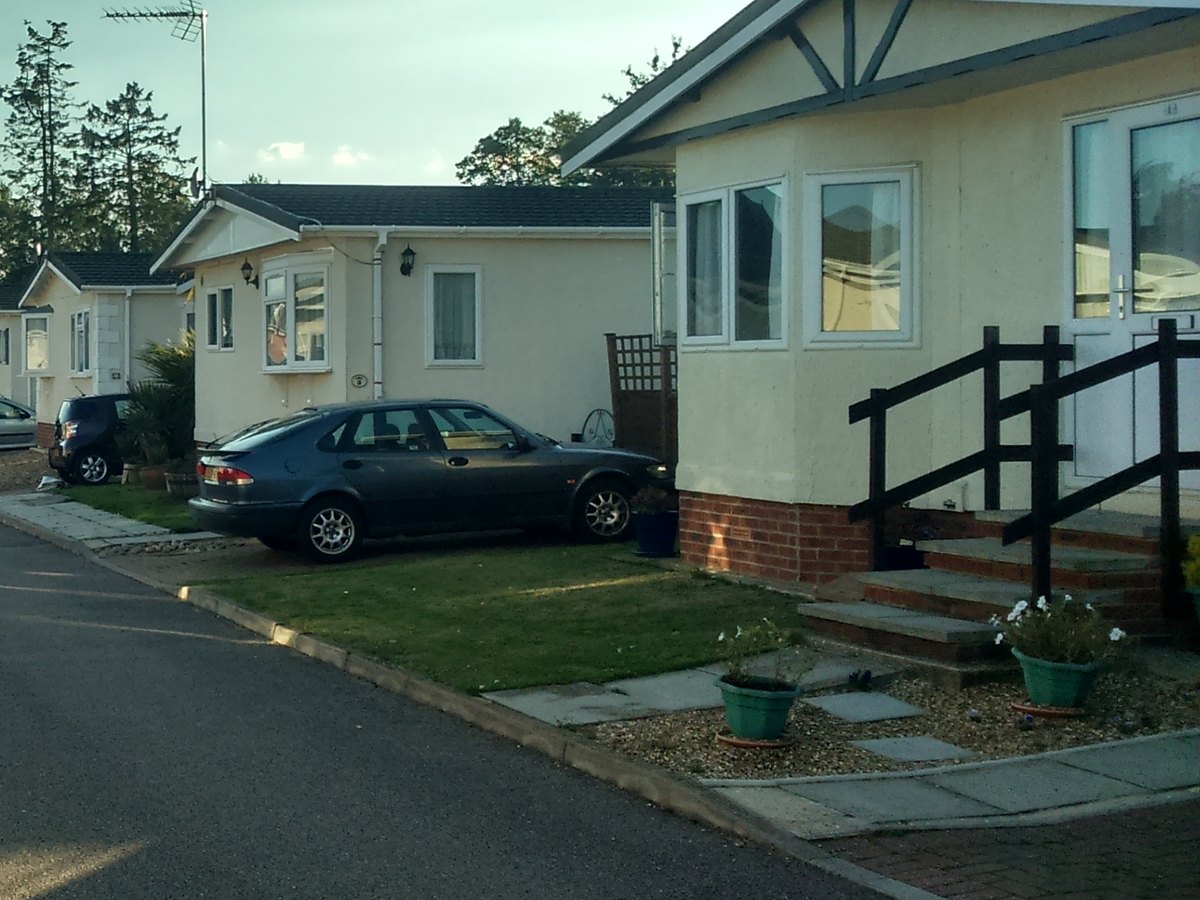 A cream park home with front steps, a handrail and flowers in pots beside a lawn